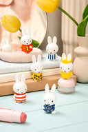 Set of small Miffy rabbit figurines in various outfits on a light surface with decorative items in the background.