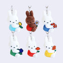6 plush keychain charms of Miffy, all holding a 2D style flat flower and sitting, facing their head to the right. 5 Miffys are white, each wearing a different color dress and 1 Miffy is brown.