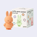 Figure of Miffy, in a soft toned pastel orange color standing in front of blind box packaging.