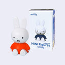 A Miffy mini figurine in its sitting pose, wearing an orange shirt, shown next to its white packaging box labeled 'miffy MINI Figures Sitting'.