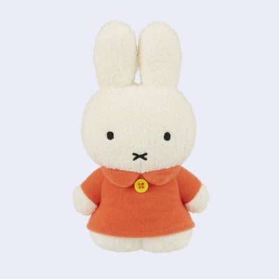 Off white Miffy plush, standing and wearing a muted orange long sleeve dress with a single yellow button around the collar. 