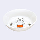 White shallow bowl with a graphic of Miffy, wearing an orange dress and standing with her hands behind her back. Small white flowers with yellow centers are around her feet.