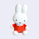 White plush Miffy, sitting in a long sleeve orange dress with a white collar and a flat yellow button.