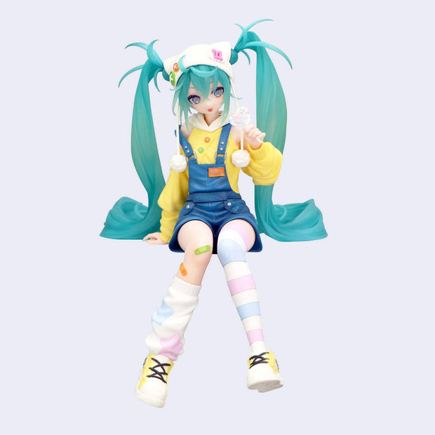 Colorful figurine of Hatsune Miku with teal hair and a yellow outfit, sitting with mismatched knee high socks and holding a pastel colored lollipop.