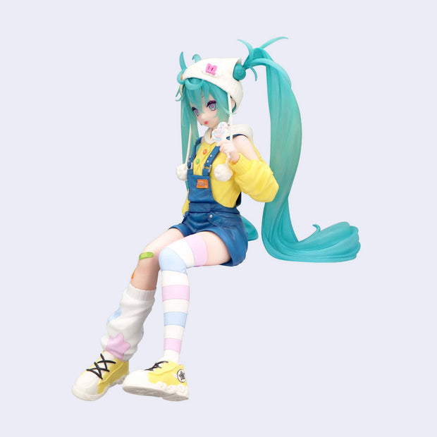 Colorful figurine of Hatsune Miku with teal hair and a yellow outfit, sitting with mismatched knee high socks and holding a pastel colored lollipop.