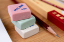 Three colorful erasers stacked on a wooden surface with a pencil and box in the background.