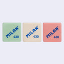 Three square erasers in green, beige, and pink with 'MILAN' branding on a light purple background.