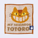 Small square brown towel with orange border and smiling orange cat face with yellow eyes and whiskers. Below is tan text that reads 'My Neighbor Totoro"