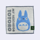 Small square towel with cream background and sideways text that reads "My Neighbor Totoro" in gray. A simple illustration of a small blue Totoro is next to it.