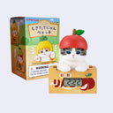 Small figurine of a gray and white cat popping up out of a fruit cartoon for apples, with a small digital clock in the front. The cat wears an apple shaped cap and sits in front of its blind box packaging.