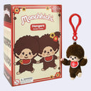 Monchhichi Exclusive Flocked Collection Hangers plush keychain shown next to its retail packaging box. The box features illustrations of the Monchhichi character, and the keychain itself is a brown plush monkey with a red heart emblem and a red plastic clip.