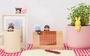 Desk setup with a mug, calendar, plant, and small figurines on a checkered tablecloth.