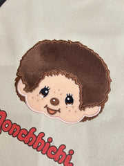 Close up of tote bag showing embroidered patch of a cartoon monkey with 'Monchhichi' branding on a beige background