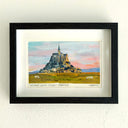 A framed artwork of Mont Saint-Michel, France, featuring a colorful painting of the abbey and landscape under a pink and blue sky, presented in a black frame with a white mat on a textured white wall.