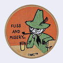 Circular embroidered sticker with Snufkin sitting at a table with a glass and text that reads "Fuss and misery.."