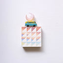 Small square wood panel painted like a pinkish blue sunset with white patterning overlayed on top, akin to a quilt tile design. Atop of it is a toy car and a carved wooden pink crescent moon.