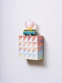 Small square wood panel painted like a pinkish blue sunset with white patterning overlayed on top, akin to a quilt tile design. Atop of it is a toy car and a carved wooden pink crescent moon.