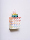 Small square wood panel painted like a pinkish blue sunset with white patterning overlayed on top, akin to a quilt tile design. Atop of it is a toy car and a carved wooden pink crescent moon.