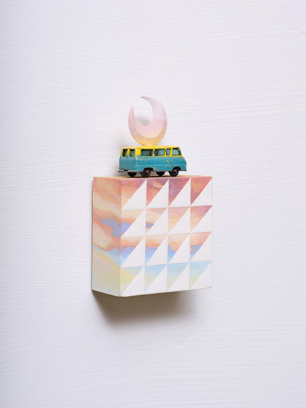 Small square wood panel painted like a pinkish blue sunset with white patterning overlayed on top, akin to a quilt tile design. Atop of it is a toy car and a carved wooden pink crescent moon.