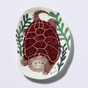 Decorative plate shaped like a slightly distorted oval with a sea turtle design and floral elements, in earthy tones of green and dark brownish red. 