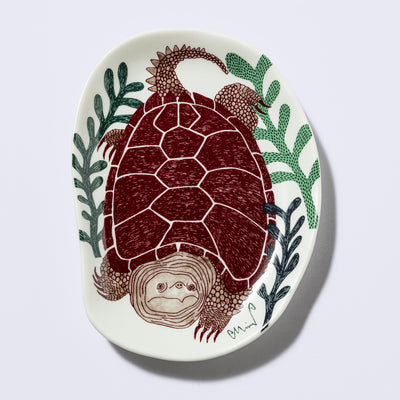 Decorative plate shaped like a slightly distorted oval with a sea turtle design and floral elements, in earthy tones of green and dark brownish red. 
