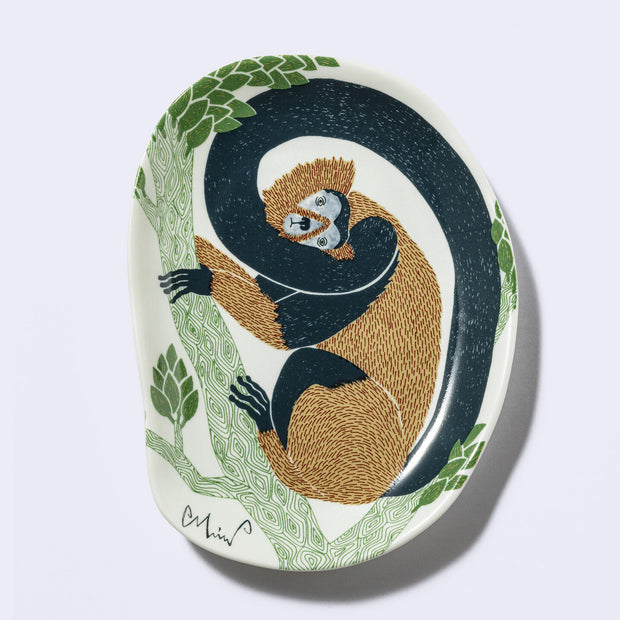 Decorative plate shaped like a slightly distorted oval with a monkey design and floral elements, in earthy tones of green, brown and dark teal. 
