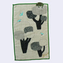 Textile art piece with tree and bird designs on a beige background. Atop one of the trees is a green bear.