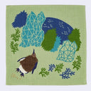 A square light green woven textile featuring a stylized nature illustration with a brown and blue bird, blue and green geometric trees, and blue-green plants.