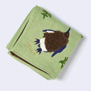 A folded sage green linen cloth towel featuring an embroidered brown bird with blue and white accents, green leaves, and gold script embroidery.