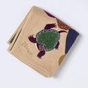 A folded beige woven towel featuring a stylized turtle design in green, burgundy, and navy blue, with a signature logo on the side, photographed on a clean white background.