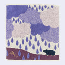 Square fabric towel with a design of many large purple rainclouds, with purple raindrops and a small hippo standing below.