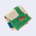 Folded square fabric towel with pink beige coloring and illustrated designs of large trunked trees and a grassy field with a small striped hyena.