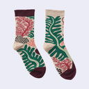 A pair of crew socks with a botanical and coral-inspired pattern, featuring green monstera leaves, red coral motifs, and dark burgundy accents on a light beige background.