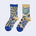 A pair of crew socks with a beige background featuring a blue and green botanical pattern, blue cuffs and toes, and a large green turtle illustration on one sock, shown against a light gray background.