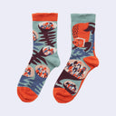 A pair of crew socks with a colorful, stylized forest and animal print. The design features a sage green background with orange, dark brown, and blue patterns of ferns, and a fox-like dog, with solid orange toes, heels, and cuffs.
