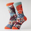 A pair of crew socks with a colorful forest pattern featuring a red-orange fox, blue leaves, and abstract shapes on a light blue background, with bright orange toes, heels, and cuffs.