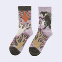 A pair of crew socks with a whimsical forest and animal print on a light lavender background. One sock features an orange tree with pink and white berries, while the other features a black wofl/badger and beige tree branches with pink berries.