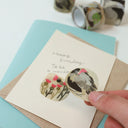 A hand holds a round sticker featuring a bird illustration over a cream birthday card with handwritten text, layered over a kraft envelope and a light blue card, with washi tape rolls blurred in the background.