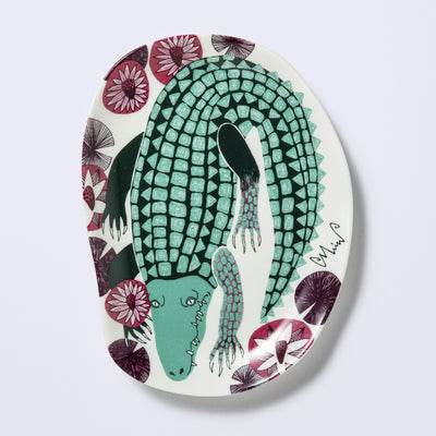 Decorative plate shaped like a slightly distorted oval with a crocodile design and floral elements, in muted tones of mint green, dark green and purplish red. 