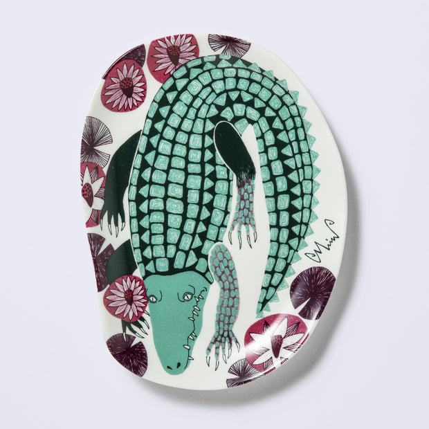 Decorative plate shaped like a slightly distorted oval with a crocodile design and floral elements, in muted tones of mint green, dark green and purplish red. 