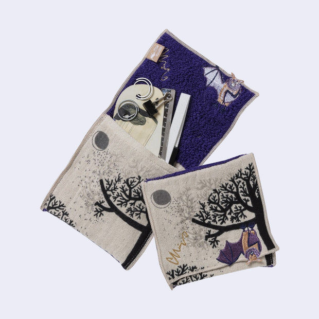 Rectangular textile with a pocket, the top can be folded over to make it into a pouch. Interior is purple and exterior is beige with designs of a purple bat, a tree and a moon. Shown both folded and with stored items within.