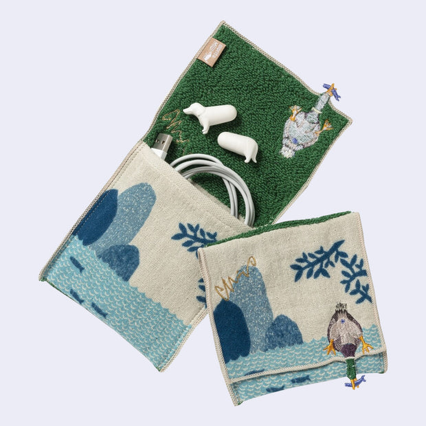 Rectangular textile with a pocket, the top can be folded over to make it into a pouch. Interior is green and exterior is beige with designs of a duck diving for a fish on water. Shown folded over and also holding items.