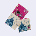 Rectangular textile with a pocket, the top can be folded over to make it into a pouch. Interior is bright fuchsia and exterior is beige with designs of a sloth hanging off of a circus tent. Shown both folded over and with items in the inner pouch.