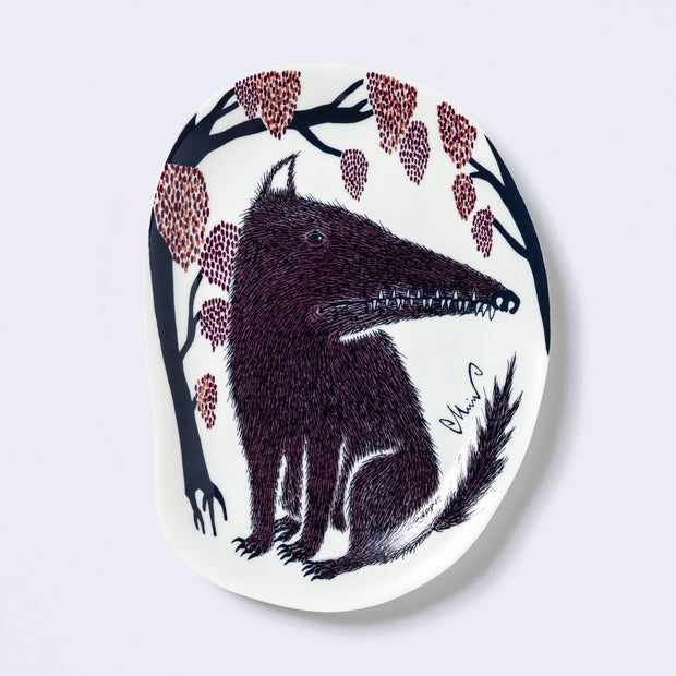 Decorative plate shaped like a slightly distorted oval with a wolf design and floral elements, in muted tones of dark blue, dark purple and dark orange/red.