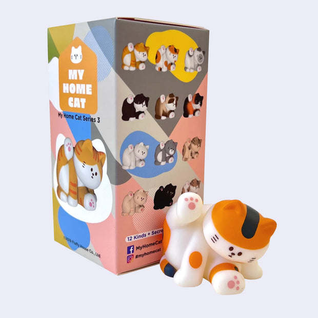 Small figurine of a calico cat, leaning down while sitting as if cleaning its underside. It sits in front of its product packaging, which shows all cat color options.