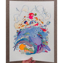 A hand holds a white art print featuring a whimsical, colorful illustration of a child in a boat surrounded by sea creatures, fish, and clouds, set against a speckled gray countertop.