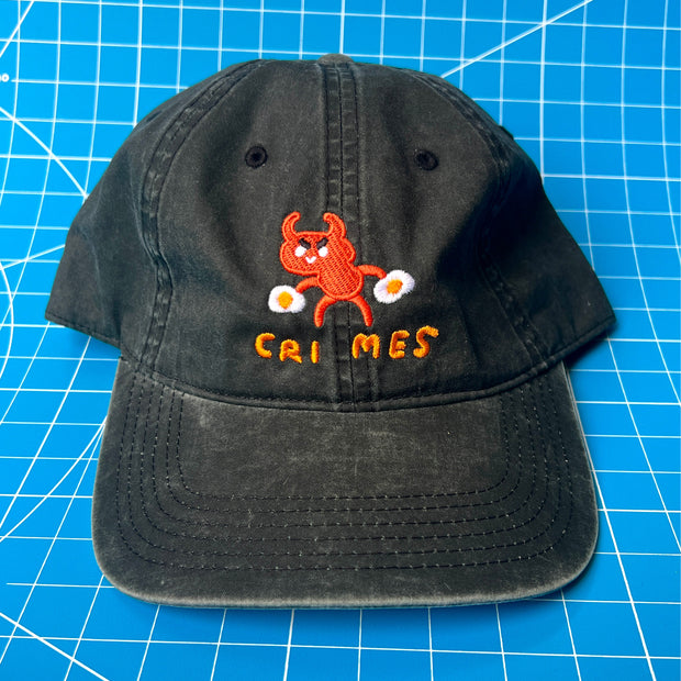 Black cap with a small embroidery in the center of a cartoon red devil with a sunny side up egg in each hand. Below it reads "crimes" which a space between "cri" and "mes."