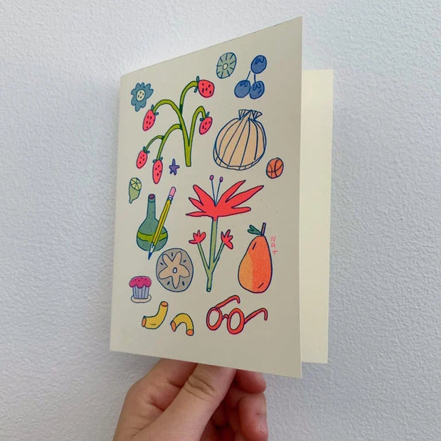Cream colored greeting card with colorful doodles on the front. Drawings include fruit, a sea shell, glasses, a pencil, vase and flowers. Card is slightly ajar showing a blank inside.