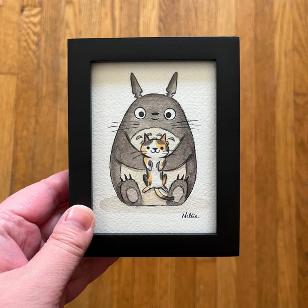 Painting of a stylized cute Totoro, sitting on the ground and holding up a calico cat in its arms. Piece is in black frame.