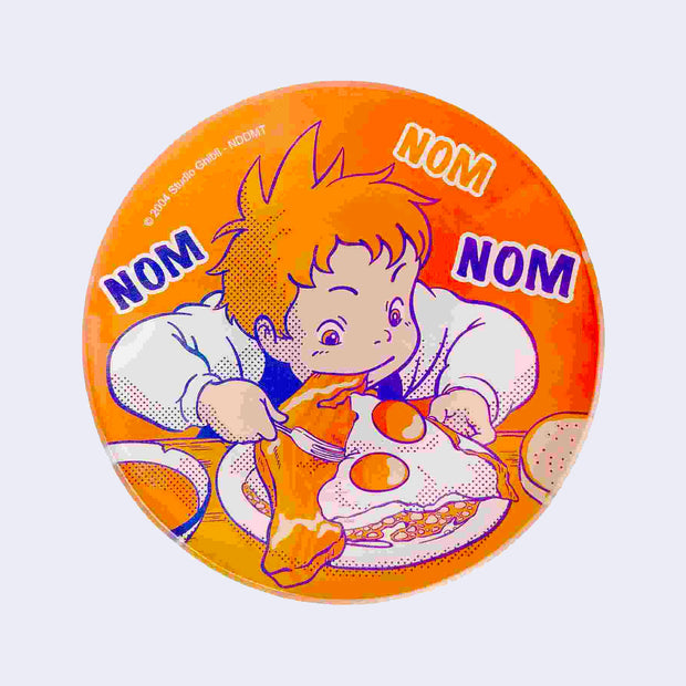 Rounded brightly colored glass plate of mainly orange and blue, featuring an illustration from Howl's Moving Castle of a young boy voraciously eating his egg and bacon breakfast. Text reads "Nom Nom Nom"